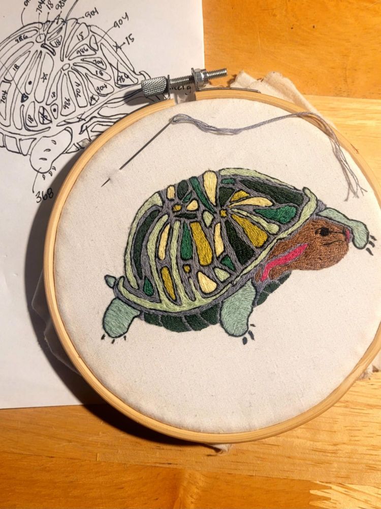 Photo of an embroidered red-ear turtle, still in an embroidery hoop with a threaded needle above the work. Behind and to the left is a black-and-white template with handwritten notes.