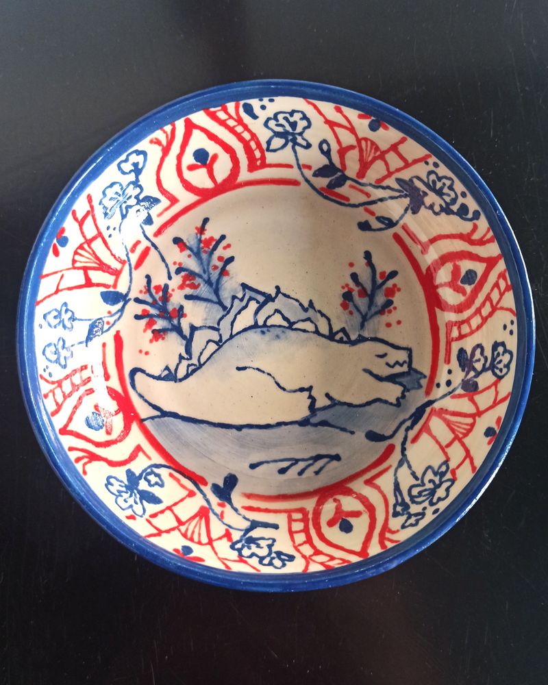 Close shot of the little ceramic dish cup depicting a small Godzilla napping.
Blue and red floral patterns on rim of the dish, framing Godzilla
Blue underglaze lineart with watercolor effects, Red underglaze patterns and details