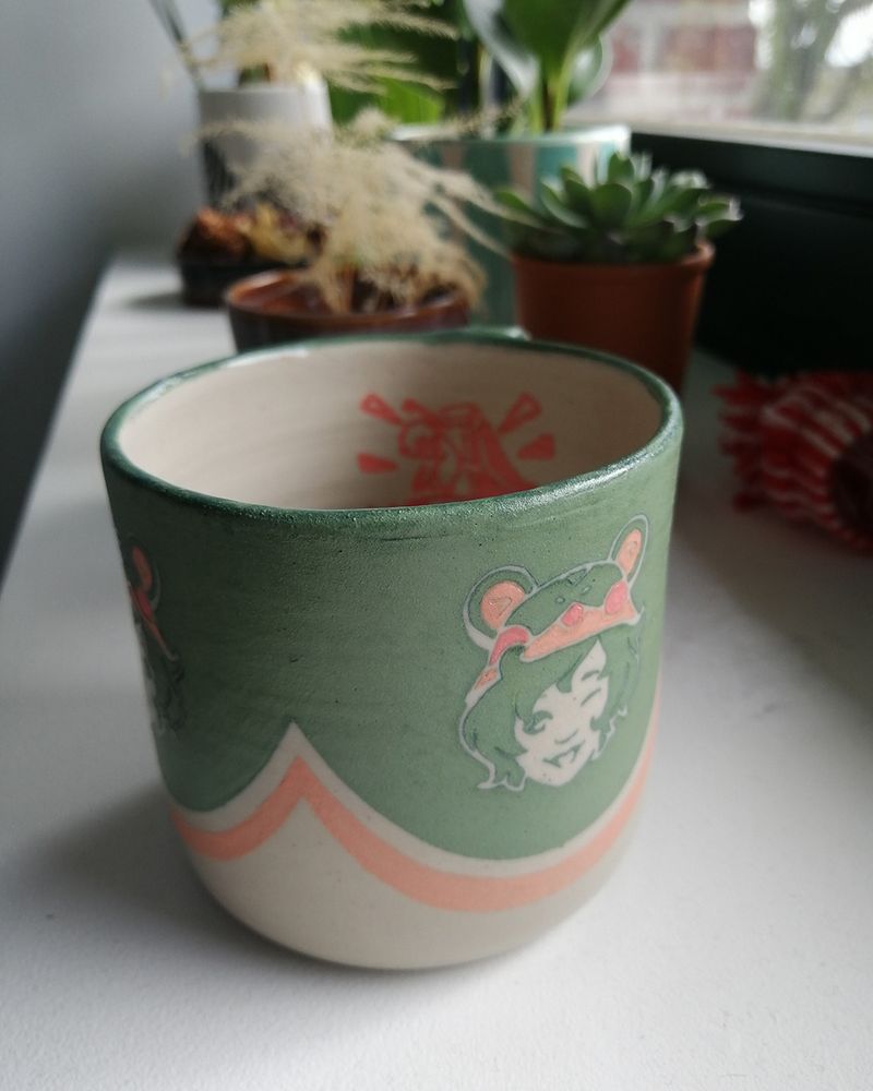 Photo of the Milio mug taken by the client, the mug is by a window , in front of several plants.
The smiling face is visible as well as the pink pattern skill in the inside of the mug