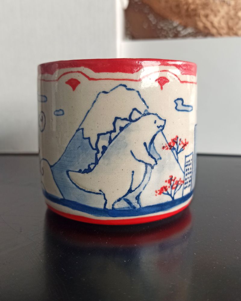 Close shot of the ceramic cup depicting a small Godzilla in front of Fujisan 
Blue underglaze lineart with watercolor effects, Red underglaze patterns and details (cherry blossom dots)