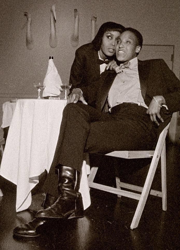 This sepia image shows two dapper individuals dressed in tuxedos sitting at a dinner table.