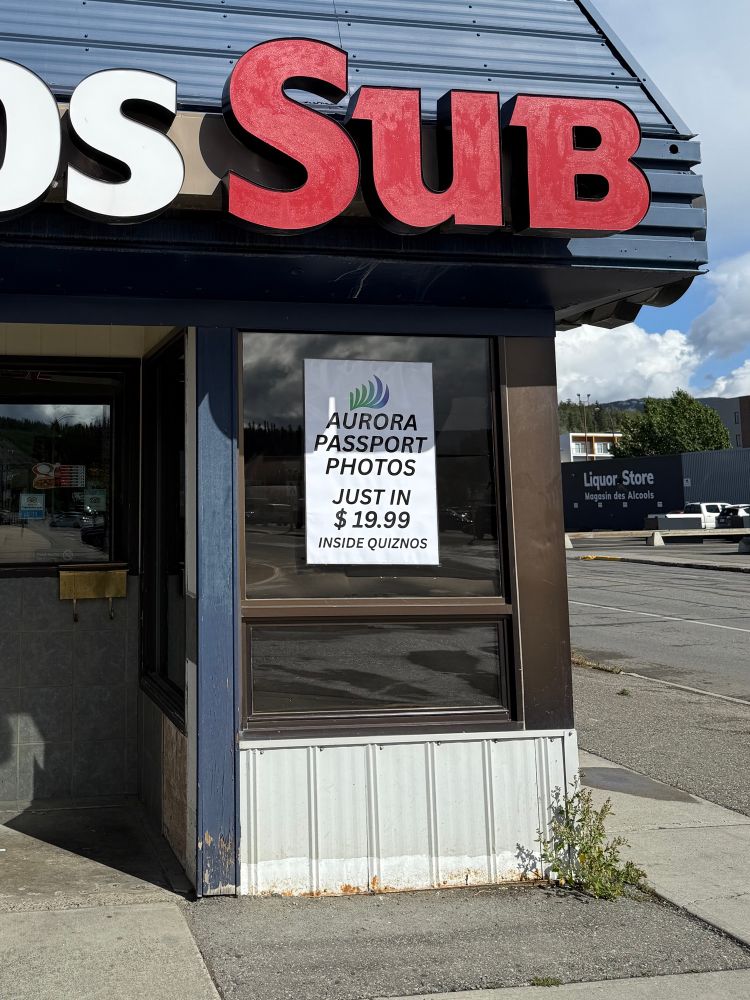 A photo of the sign in the window of Quiznos that says “Aurora passport photos. Just in $19.99. Inside Quiznos.”