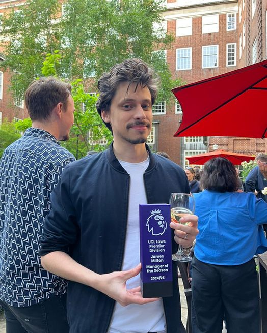 James is holding a glass of wine and a purple Premier League award stands outdoors at a social gathering, with people and red umbrellas in the background.