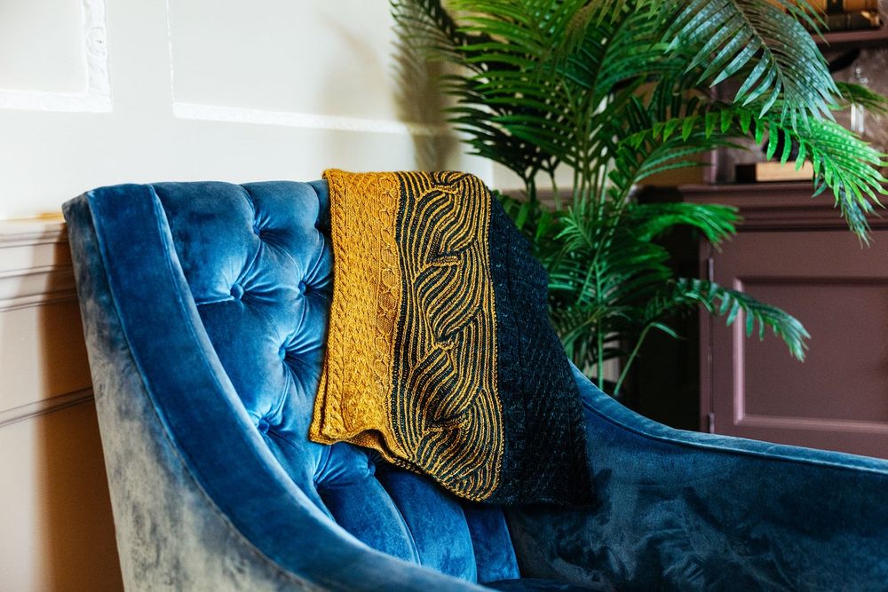 A black and gold knitted shawl draped over a blue velvet armchair, with a green tropical plant behind