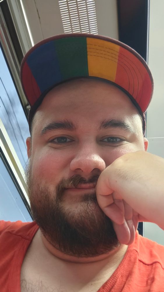 A photo of me, a bearded man, smiling into the camera. Wearing a rainbow striped hat and an orange shirt. 
