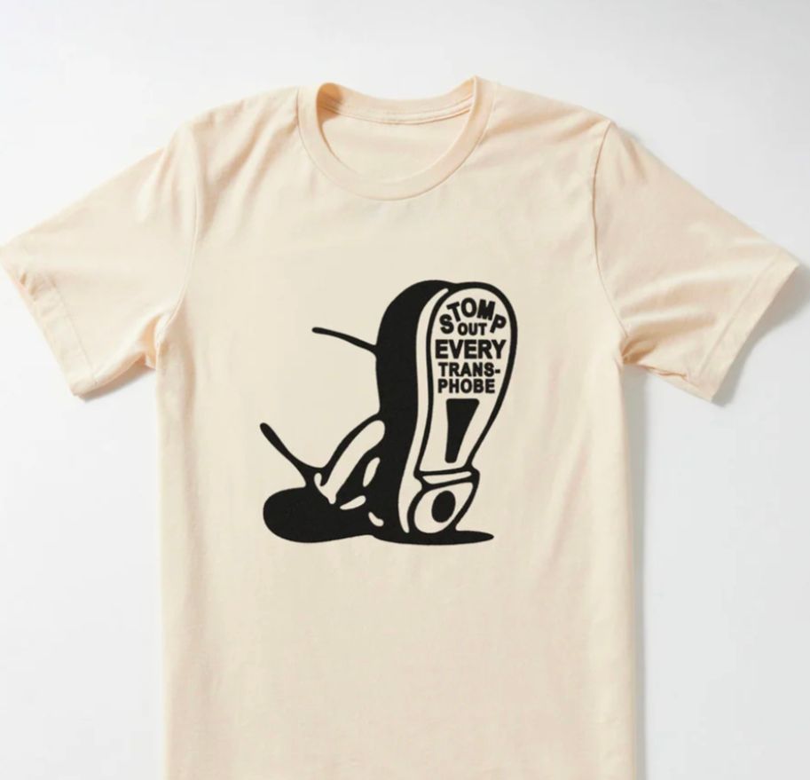 A cream colored t-shirt with an illustration of a foot and on the sole of the shoe it reads "stomp out every transphobe"