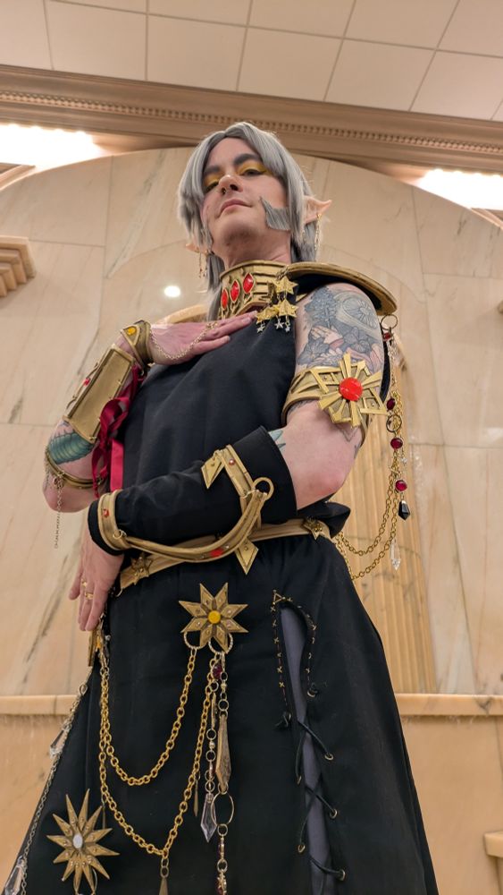 Photo of urianger cosplay from a below angle, I stand with one hand on my chest and the other crossed over my stomach