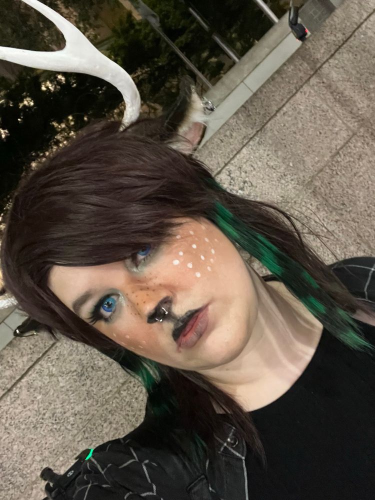me cosplaying the character december holiday from deltarune – i’ve got deer antlers and ears (which are pierced), deer makeup, and a dark brown wig styled to be “scene” with green and black raccoon stripes on either side of my face