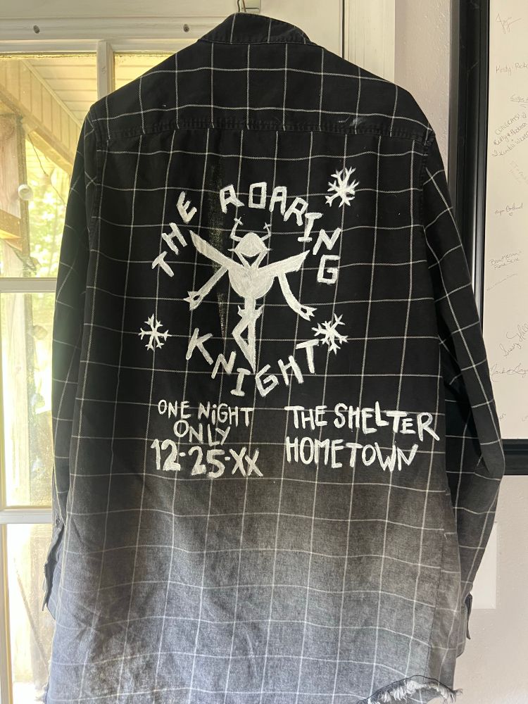 the back of the flannel. it has painted on it in white  “the roaring knight / one night only 12-25-XX / the shelter, hometown”

there is a depiction of the knight from deltarune in the middle of the text and some stylized snowflakes around it
