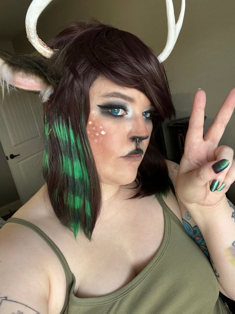 a selfie of me in cosplay deer makeup with a scene-styled wig, deer antlers and horns. i’m wearing an lil e green ribbed spaghetti strap top and am holding up a peace sign