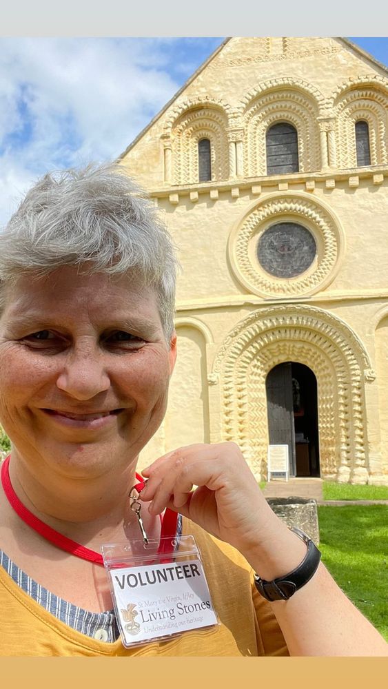 smiling woman holding up a volunteer's badge in front of the west end of a Norman church in sunshine
