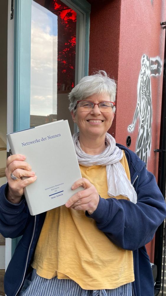 Grinning woman holding a fat grey book in front of a house door with a skeleton graffiti