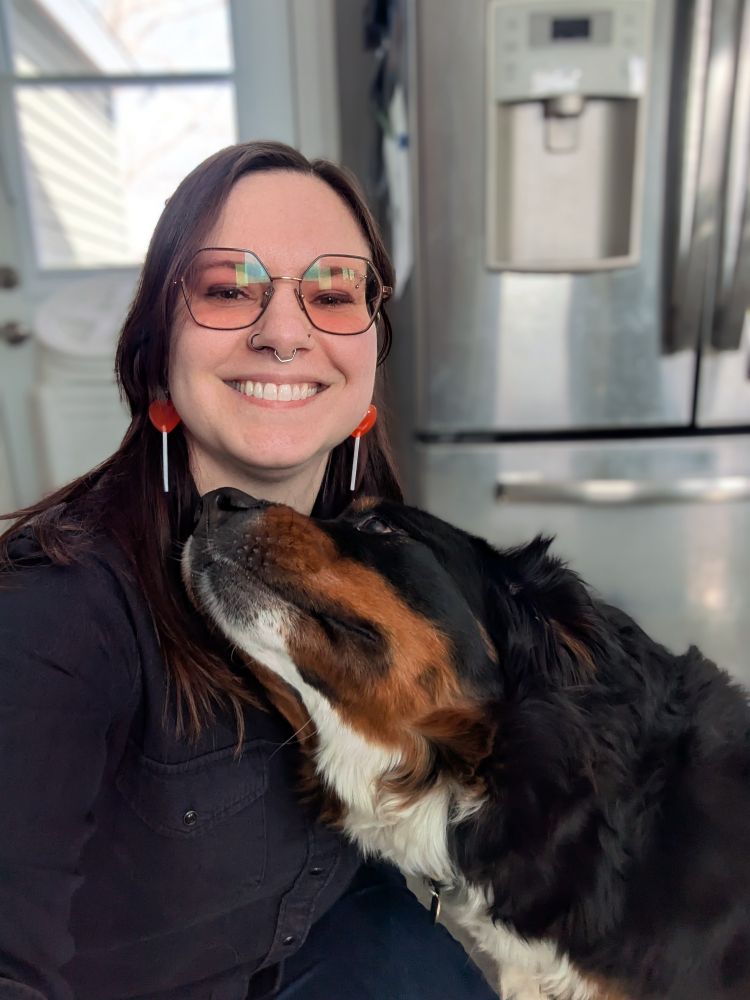 A selfie, wearing earrings that are heart sucks, a Bernese mountain dog pressed against the person's chest looking up at them