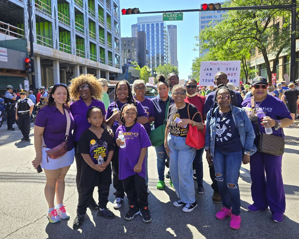 A groups of adults and children in purple SEIU shirts pose at an outdoor rally