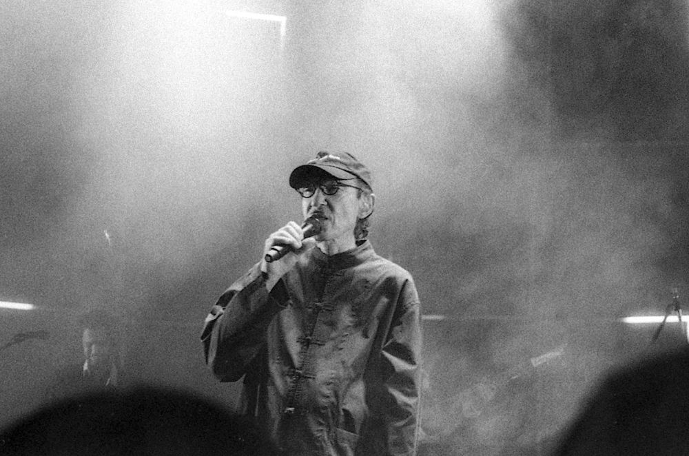 A slightly grainy black & white film photograph, taken at Hammersmith Apollo, of the legend, Ron Mael of the band Sparks, microphone in hand, during his performance of Suburban Homeboy. He is baggy enough for us.