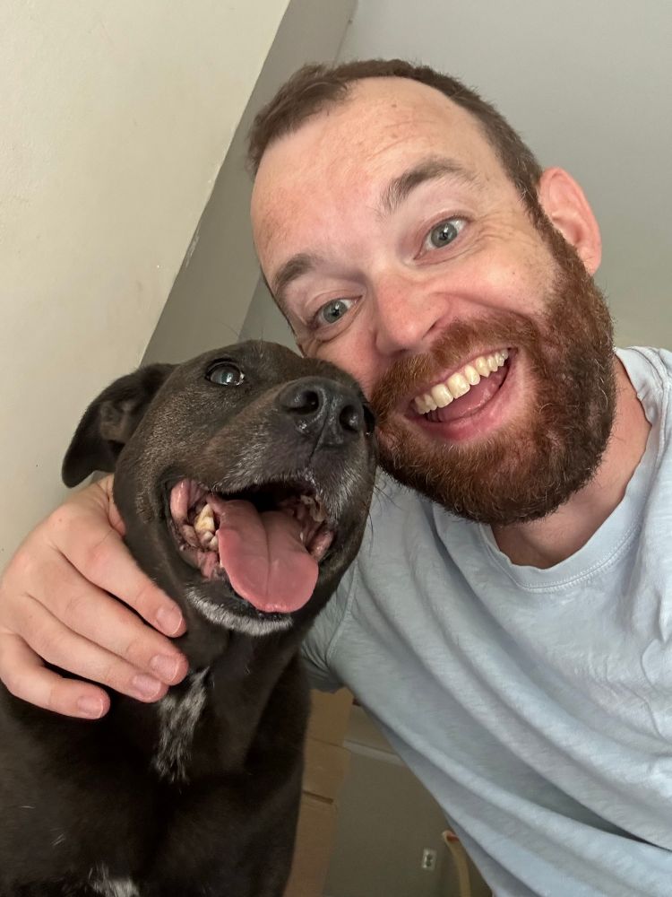 Nick, a bearded man in his forties, smiles and hugs Goose, a small black dog of indeterminate breed, who also smiles. 