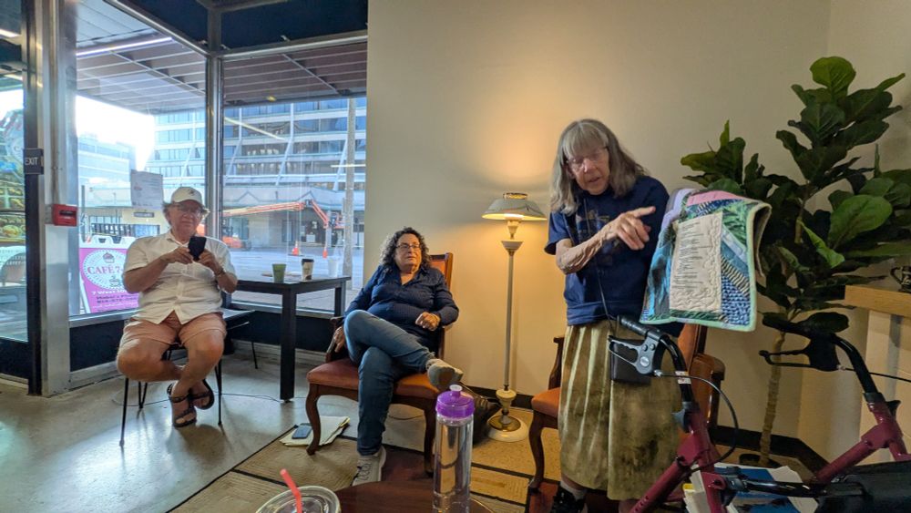 Elder femme with quilt reads a poem on it in a cafe