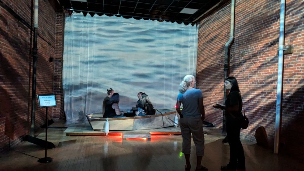 Group of six sitting in and standing around an old boat in a gallery with video projections of water