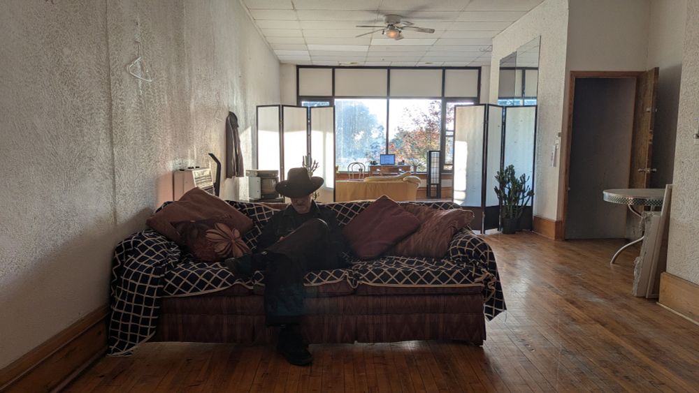 Silhouette masc dressed in black sits on a couch in a minimally furnished loft apartment with wood floors