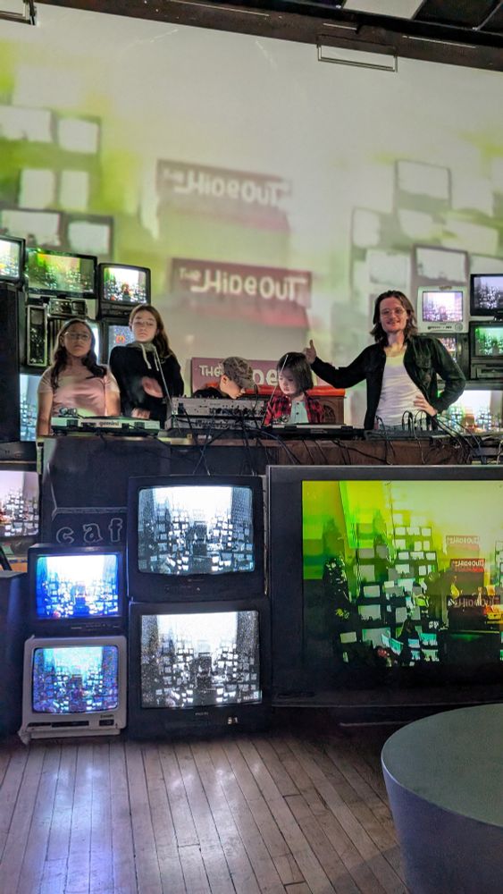 A group of children and one adult seen in a stack of Tube TVs displaying video feedback of the same scene