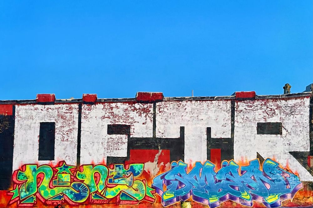 Detail of a large graffiti mural on an exterior brick wall reads OFTR NINE and SMEW, seen under bright summer sun in a cloudless blue sky 