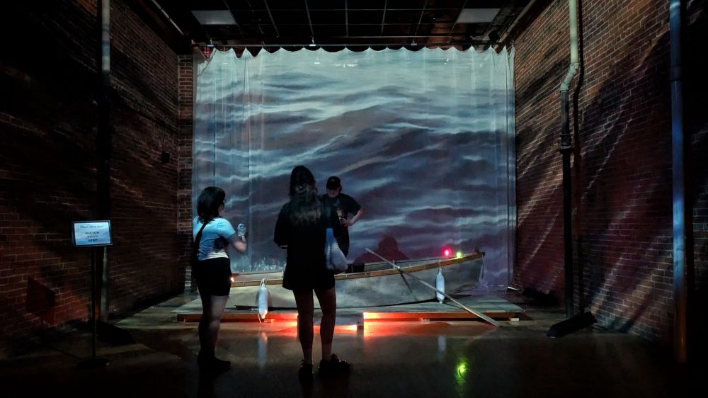 Group of 3 stands around an old small boat in a gallery with projections of lake waves illuminating the scene