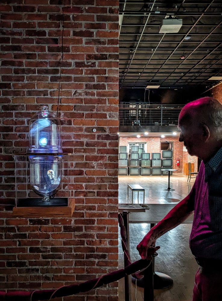 Elder masc gazes at an art exhibition of two glass bowls with projections of skull and eyeball floating inside. In the background are stacks of tube TVs 