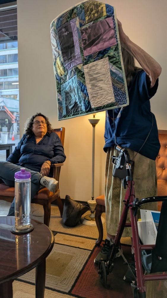 Figure holds up a quilt with  colorful patterns and a poem