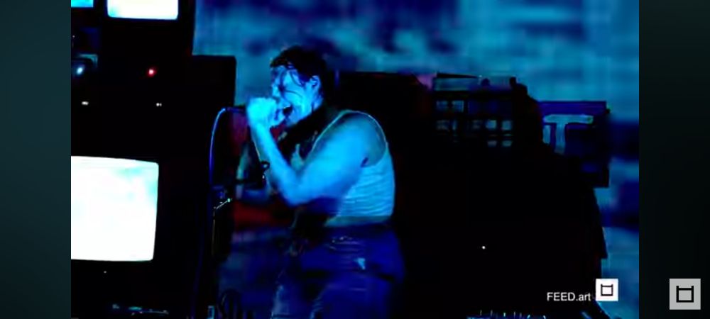 Singer screams into mic in blue projections and surrounded by Tube TVs 
