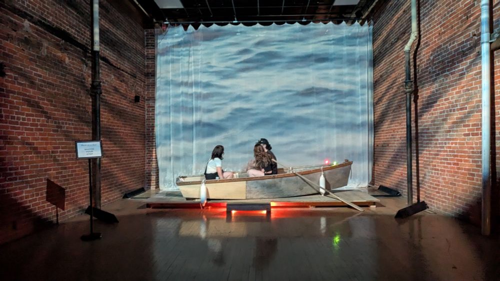 Trio sit in a small old wooden boat in a brick hall, immersed in projections and gentle waves on a lake