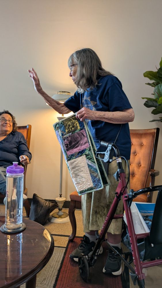 Elder femme with a walker and quilt gestures while speaking to listener in a cafe