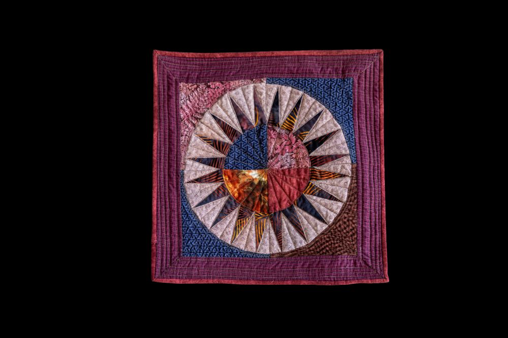 Square quilt in purples, blues, oranges, yellows with a center circular pattern divided into quadrants and rays emanating