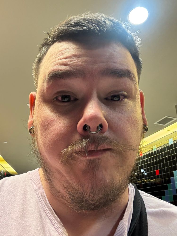 a selfie of me after watching Wicked: For Good. my eyes are bloodshot from crying. I have a septum piercing in and two piercings on my ear lobes. I have a lavender shirt on and you can see the strap of my messenger bag.