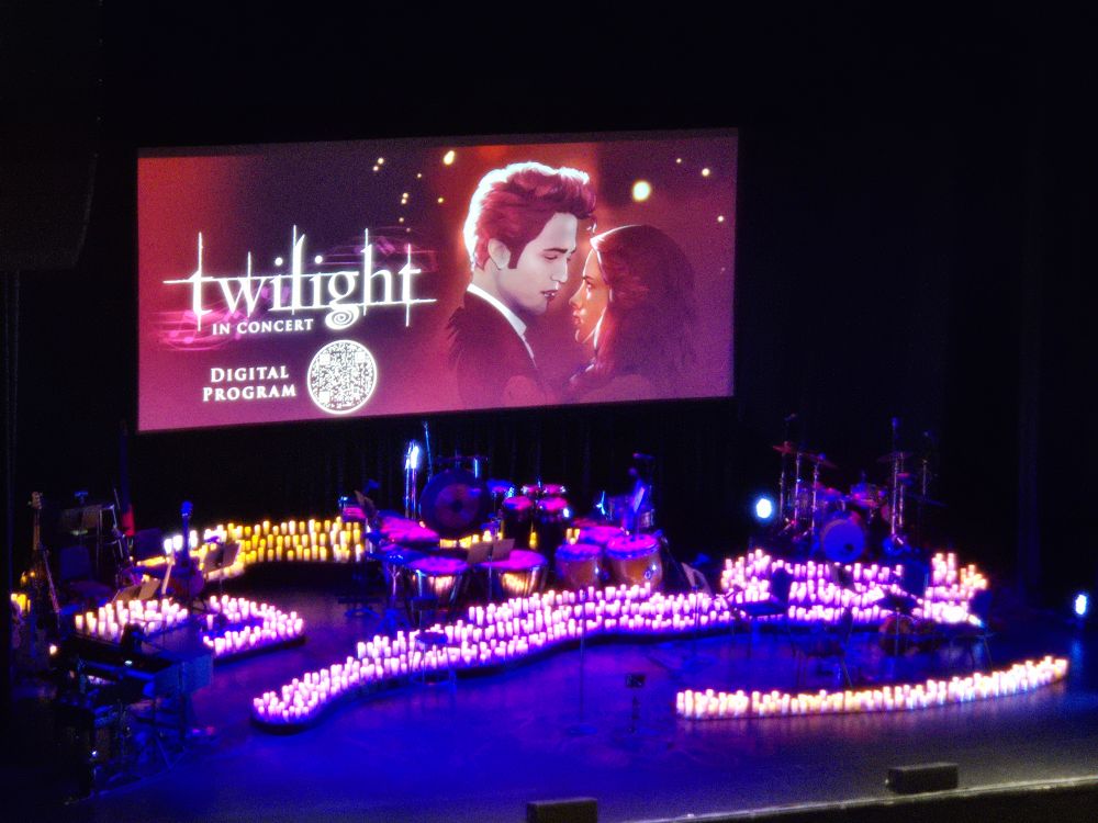picture from twilight in concert. a screen portrays edward and bella from twilight, while hundreds of candles light a stage with musical instruments below the screen. 