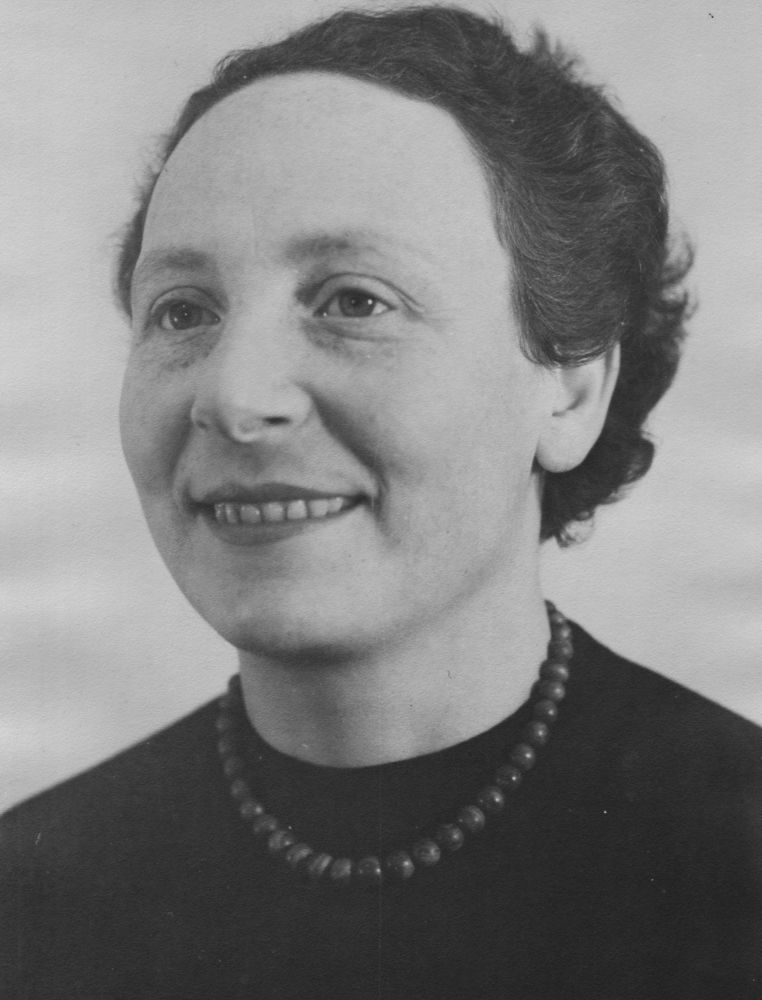 A black and white close-up portrait of a woman in her 40s, smiling warmly, looking to the side. She wears a natural look, with short, wavy hair, a black jumper, and a beaded necklace.