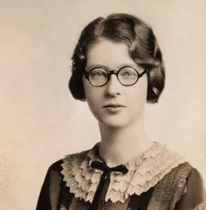 A sepia-toned portrait of a young white woman with short wavy hair and stylish, round glasses. She looks serious. Plain white background.