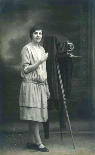 A black and white full body portrait of Karimeh Abbud standing with pride next to her professional camera. She has short, dark hair, and a light colour plain modern dress.