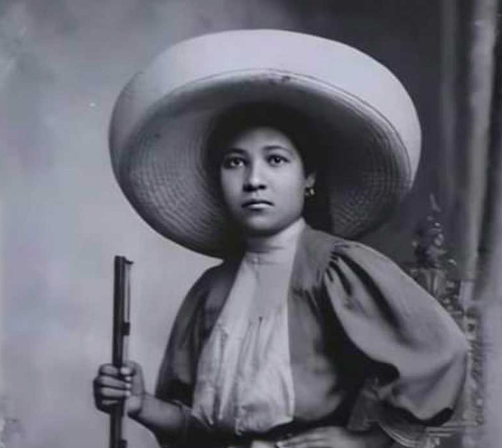 A black and white photo of a young Mexican woman wearing a big sombrero, a nice dress with wide sleeves, and she is holding a rifle. She looks serious.