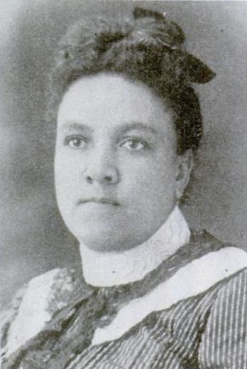 A black and white close up portrait of a young Black woman. She has a contemplative expression, with her hair neatly styled and adorned with a bow. She is dressed in period clothing, likely from the late 19th or early 20th century, featuring a high-collared blouse adorned with dark lace.