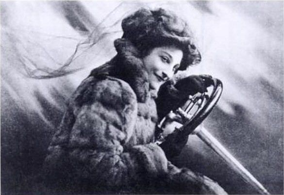 Black and white photo of Dorothy Levitt, a young white woman wearing a luxurious fur coat, smiling while holding the car's steering wheel. Her smile tells us that she really loves cars.