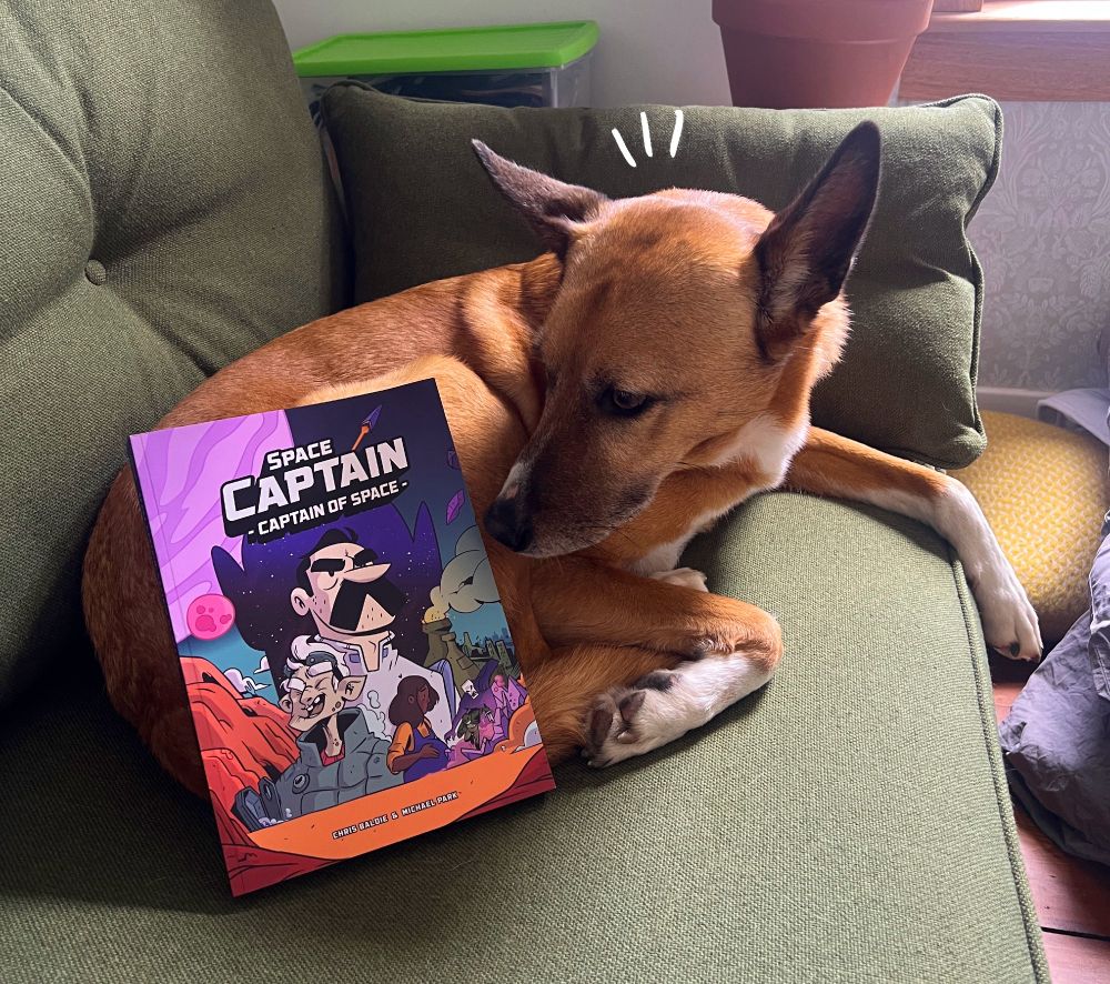 Noodle the dog, the world prominent scholar on Space Captain, is sat on a green couch with a copy of the new Space Captain book propped up on her button. She is sniffing it and from her expression it smells great. 