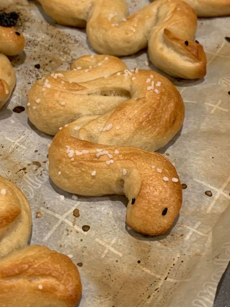 photo of a snake shaped soft pretzel on the baking sheet with other nearby snake pretzels. it has black sesame eyes