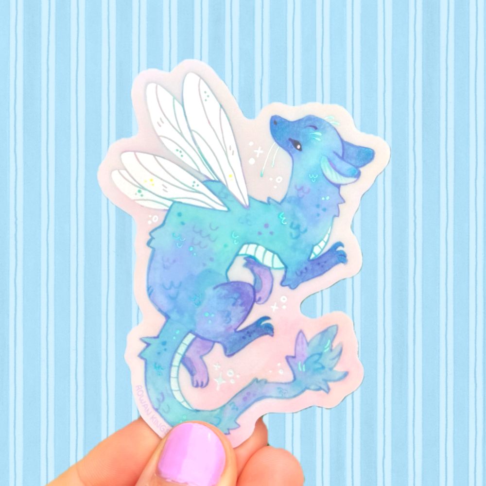 A hand holding up a sticker of a fairy dragon