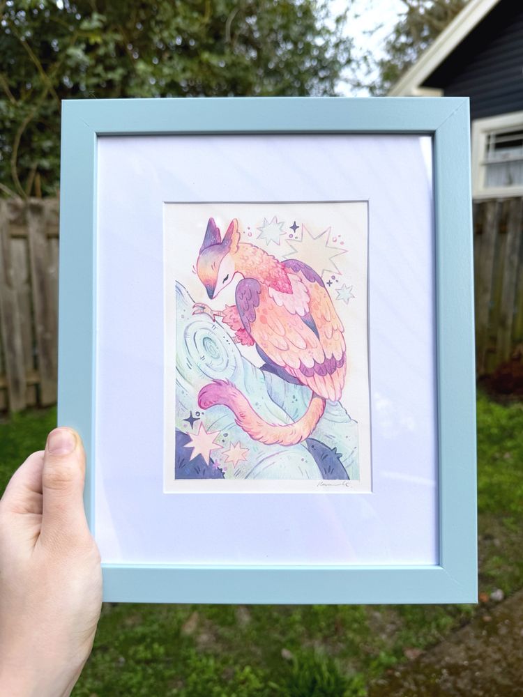 A hand holding up an original framed painting of a gryphon