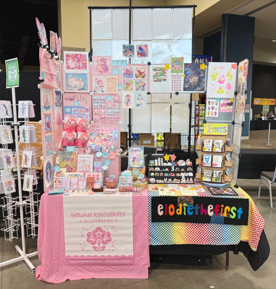 Picture of an artist alley booth. One half is pink, the other is rainbow themed. 