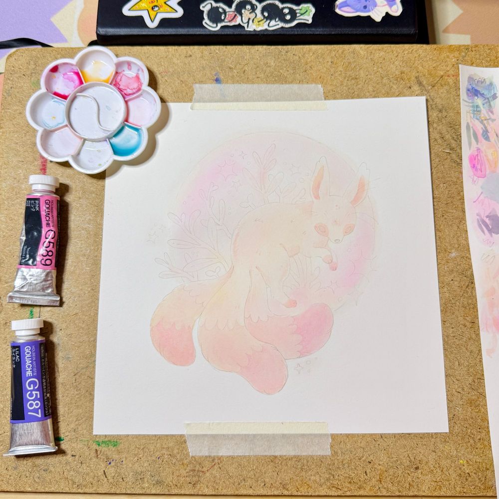 A tabletop with painting supplies and a piece of watercolor. The base layer of orange paint has been laid down on the painting.