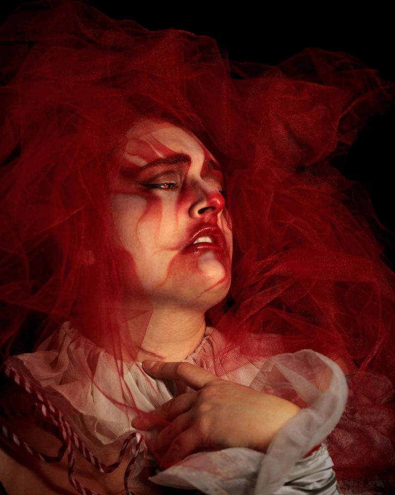 Moody expressive self portrait, with red clown inspired makeup and soft white fabric ruffles around their neck and wrist. Red flowy fabric around their head fills the frame. The subject looks to the side, bringing their hand up towards the surgical scar on their neck.