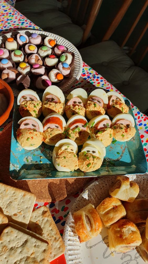 12 terrifying looking party snacks made from eggs and hotdogs to look like babies. Surrounded by other plates with sausage rolls and crackers and marshmallows.