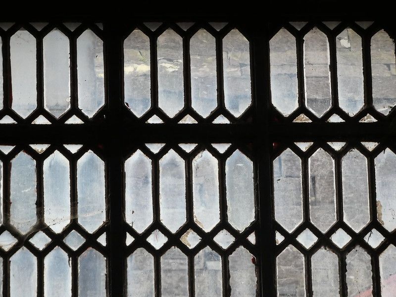 A window in the Wales Slate Museum in Llanberis. It has two thick upright iron bars, crossed by one thick horizontal bar, with smaller lattice panes of diamond and pointed rectangle panes of glass. The glass is dirty, obscuring the view of a building outside. The windows were all made on site and used small panes of glass in order to prevent the windows breaking during blasting in the nearby quarries.