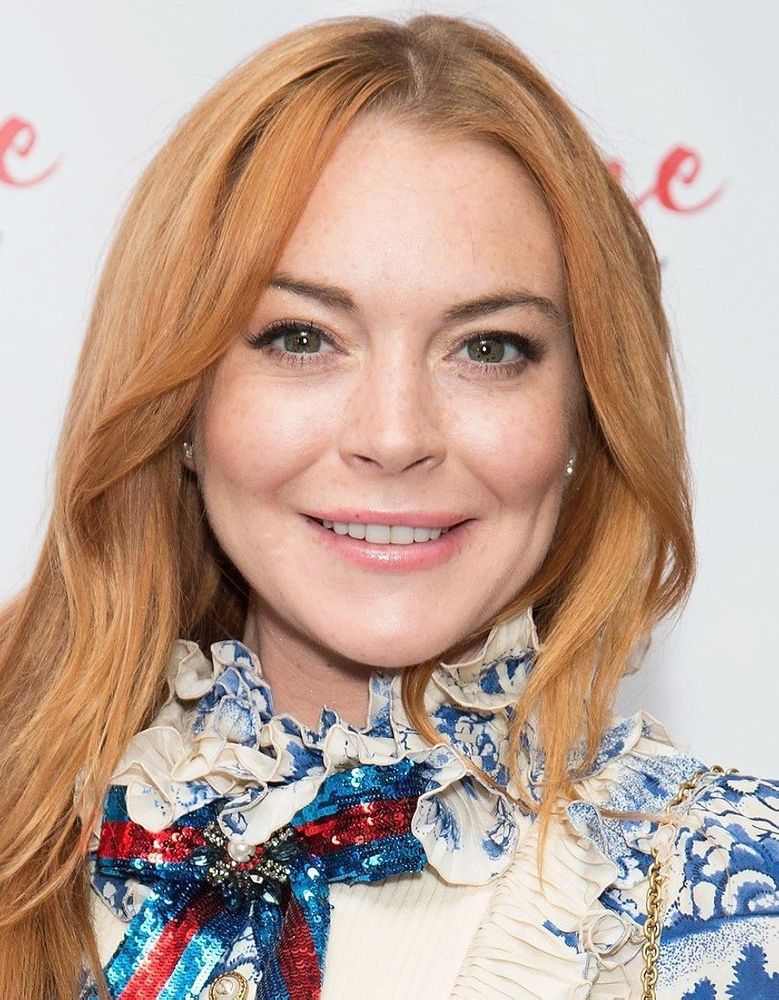A straight-on photo of Lindsay Lohan.  She has red hair of many hues that rests on her left shoulder while cascading out of view on her right side.  She smiles, showing her teeth, and her dimples accent her soft cheeks.  She wears a white blouse embellished with blue nature patterns.  A red and blue sequined, bow-shaped brocade adorns her center buttons.  She stands against a white background with obtuse red written script.